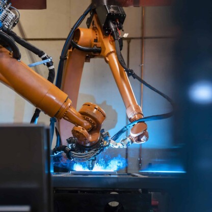 Two orange industrial robotic arms perform automated welding on a metal surface, producing blue sparks inside a factory setting—far from the quiet focus of law offices where Chicago lawyers handle complex cases.