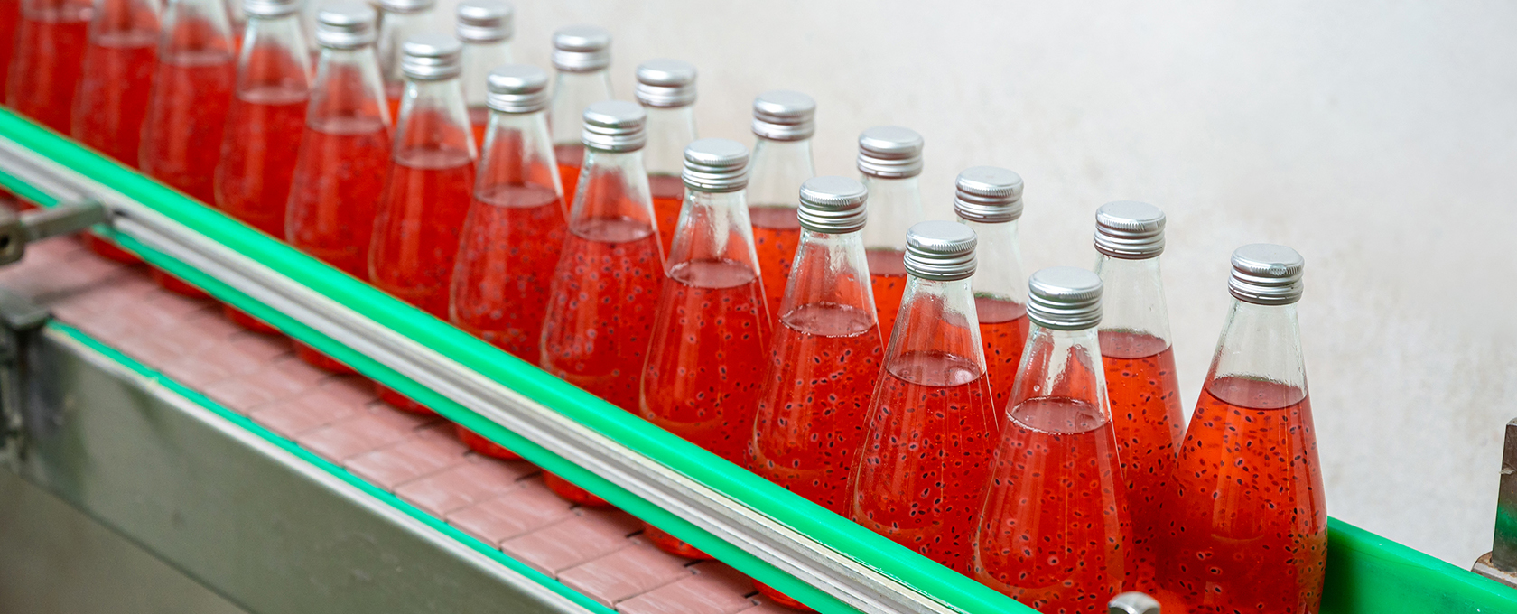 Glass bottles filled with a red beverage containing visible seeds move along a conveyor belt in a bottling facility, overseen by experts from a corporate law office.