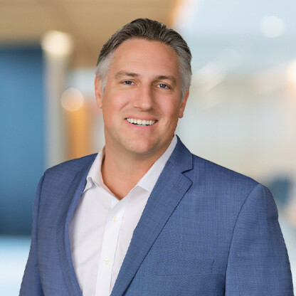 A man in a blue suit jacket and white shirt smiles while standing in a modern, blurred corporate law office.