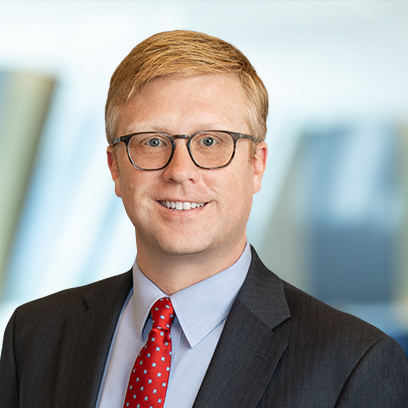 A man with short blond hair and glasses wearing a dark suit, light blue shirt, and red polka dot tie is pictured against a blurred corporate law office background.