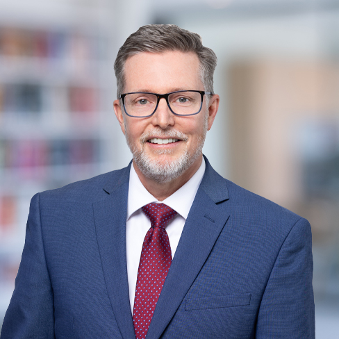 A man wearing glasses, a blue suit, white shirt, and red tie, smiles while standing in an office setting with blurred bookshelves in the background—typical of Chicago lawyers specializing in intellectual property law.