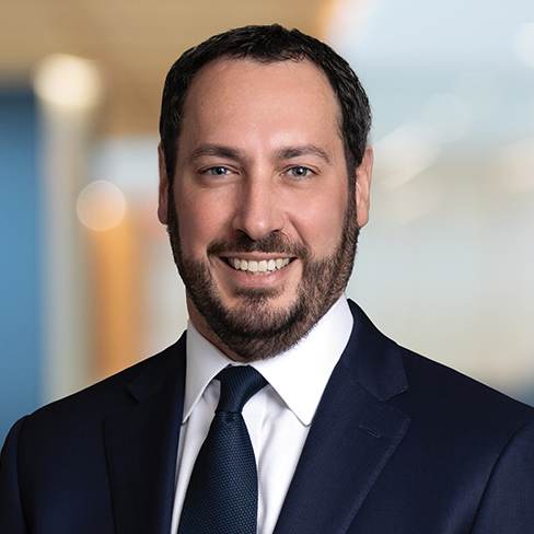 A man with short dark hair and a beard, wearing a navy suit, white shirt, and dark tie, smiles at the camera in a corporate law office setting.