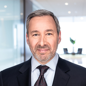 A middle-aged man with short gray hair and a beard, wearing a suit and tie, poses in a modern law office setting with glass walls—perfect for Chicago lawyers specializing in intellectual property law.