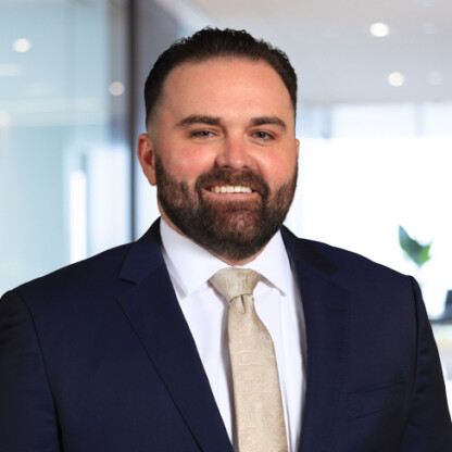 A man with short dark hair and a beard wearing a navy suit, white shirt, and patterned tie stands in a modern corporate law office.
