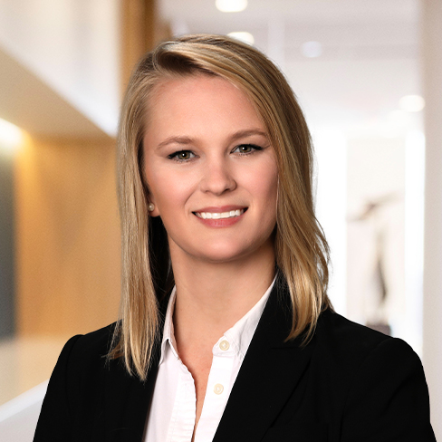 A woman with straight blonde hair wearing a black blazer and white shirt, smiling in a modern, well-lit corporate law office.