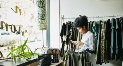 A person stands in a clothing store, looking at their phone while holding a pair of pants, perhaps searching for lawyers in Chicago, with racks of clothing and a mannequin visible in the background.
