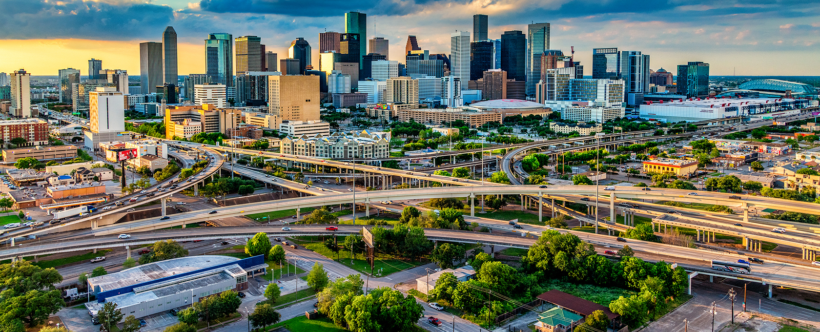 Aerial view of a city skyline with tall buildings, highways, and overpasses intersecting in the foreground—among them, prominent law offices providing litigation support—under a partly cloudy sky at sunset.
