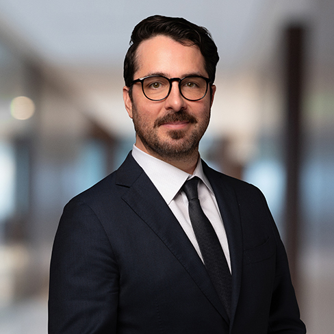 A man in a dark suit, white shirt, and black tie stands in a modern, blurred corporate law office hallway, looking at the camera.