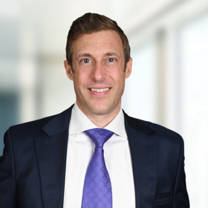 Man wearing a dark suit, white shirt, and purple tie stands in front of a blurred corporate law office background, smiling at the camera.