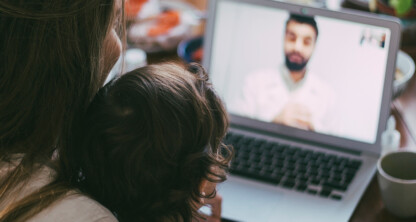 A woman and a child sit together facing a laptop, participating in a video call with a man from a corporate law office.