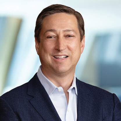 A man in a navy suit and light blue shirt smiles at the camera with a blurred corporate law office in the background.