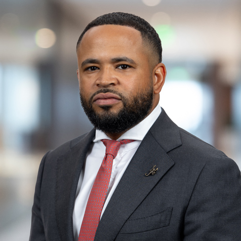 A man with a trimmed beard and short hair, dressed in a dark suit, white shirt, and red tie, stands confidently in the blurred backdrop of a corporate law office.
