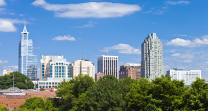 Raleigh city skyline with modern high-rise buildings, including a prominent corporate law office, and trees in the foreground under a blue sky with scattered clouds.