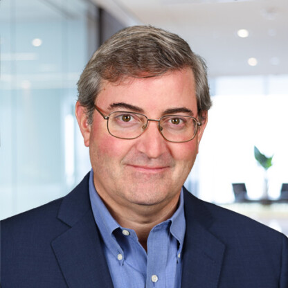A middle-aged man with gray hair and glasses, wearing a blue suit jacket and blue shirt, stands in a modern corporate law office—an ideal setting for experienced lawyers in Chicago.