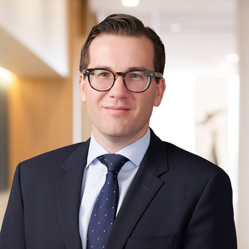 A man wearing glasses, a navy suit, and a blue polka-dot tie stands in the well-lit hallway of a modern corporate law office.