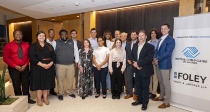 A group of people pose indoors next to a Boys & Girls Clubs of America and Foley & Lardner LLP banner at a corporate law office.