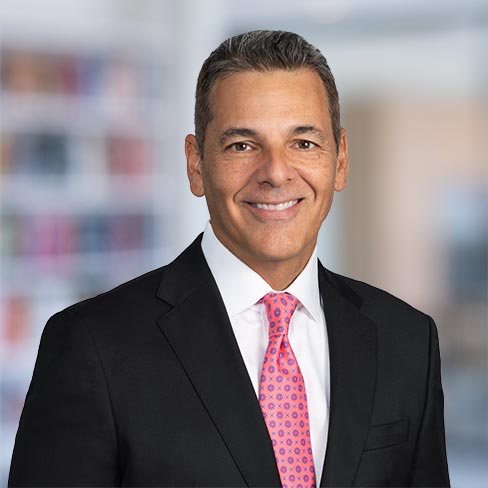A man in a black suit, white shirt, and pink patterned tie smiles at the camera with a blurred office background, representing the professionalism found at leading law offices and among top Chicago lawyers.