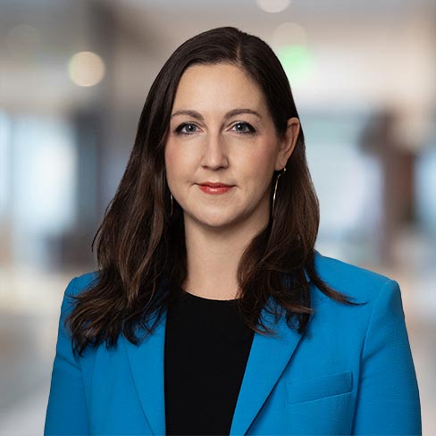 A woman with long brown hair wearing a blue blazer over a black top, standing in a corporate law office with a blurred background, representing chicago lawyers.