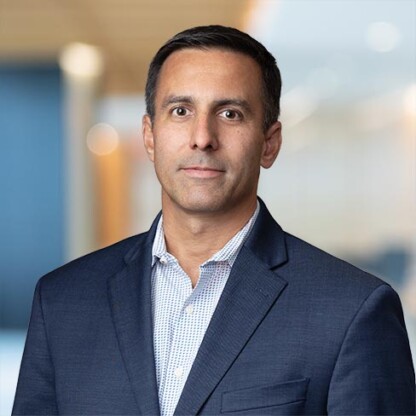 A man in a navy blazer and light patterned shirt stands in an office setting, with a blurred blue and beige background—a professional scene often found in law offices providing top-tier litigation support.