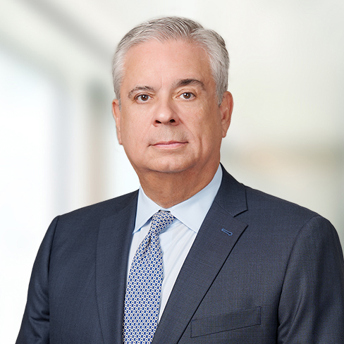 A middle-aged man with gray hair wearing a dark suit, light blue shirt, and patterned tie stands confidently in front of a blurred corporate law office background.