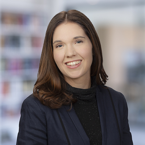 A woman with long brown hair wearing a dark blazer and black top smiles at the camera against a blurred corporate law office background, representing professional lawyers in Chicago.