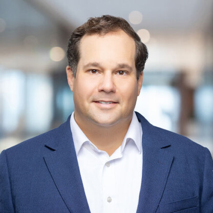 A man with short brown hair, wearing a white shirt and blue blazer, stands in a brightly lit indoor setting with a blurred background, evoking the professional image of chicago lawyers in modern law offices.