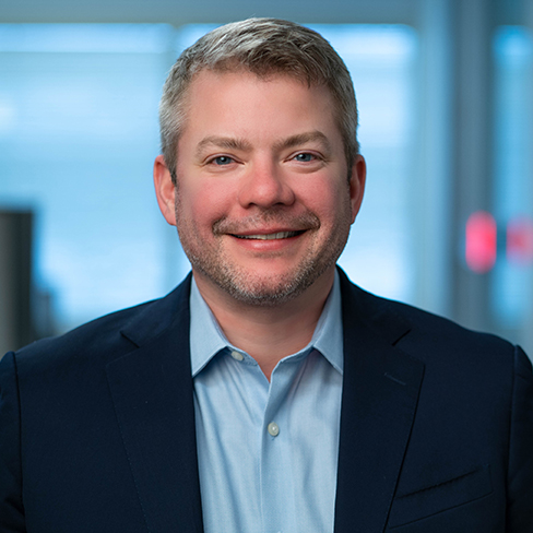 A man with short gray hair and a trimmed beard, wearing a blue suit jacket and light blue shirt, smiles at the camera in a law office—a professional dedicated to providing litigation support for Chicago lawyers.