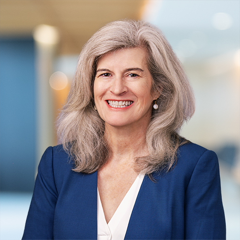 A woman with long gray hair wearing a blue blazer and white blouse smiles at the camera in a modern corporate law office, reflecting the professional environment of lawyers in Chicago.