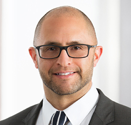 A man with glasses and a beard wearing a suit and striped tie, standing in front of a bright, neutral background—exuding the professionalism often seen among Chicago lawyers specializing in intellectual property law.