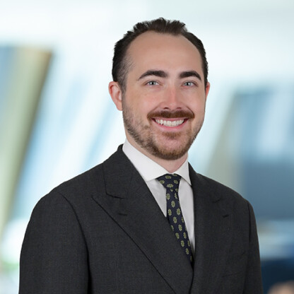 A man in a dark suit, white shirt, and patterned tie smiles at the camera against a blurred law offices background.