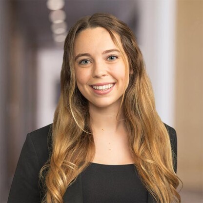 Woman with long wavy blonde hair wearing a black blazer and top, smiling, standing in a hallway with blurred background of law offices.