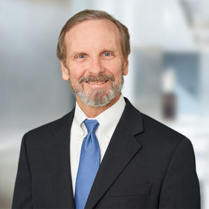 A middle-aged man with a beard and mustache, wearing a dark suit, white shirt, and blue tie, stands in front of a blurred corporate law office background, smiling at the camera.