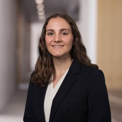 A woman with long brown hair in a dark blazer and light blouse stands in a blurred indoor hallway, facing the camera and smiling—a confident professional among Chicago lawyers specializing in intellectual property law.