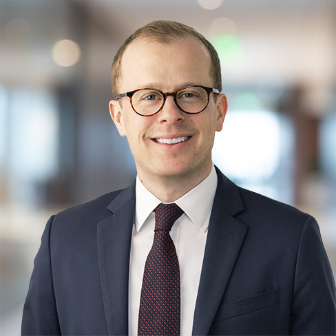 A man wearing glasses, a navy suit, white shirt, and patterned tie smiles while standing in a brightly lit, blurred office hallway—typical of successful Chicago lawyers.