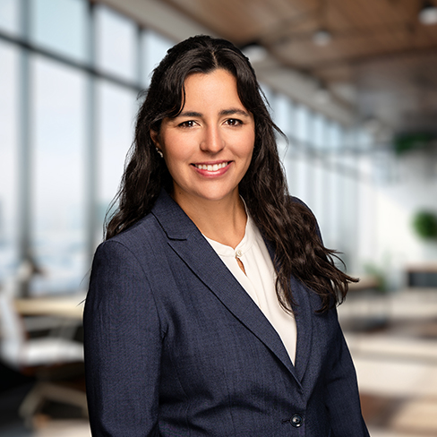 Woman with long dark hair, wearing a navy blazer and white blouse, smiles while standing in a modern corporate law office in Chicago with large windows and bright natural light.