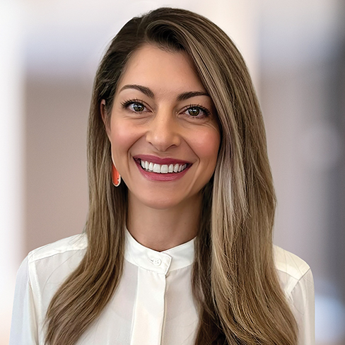 A woman with long, straight light brown hair, wearing a white blouse and orange earrings, smiles at the camera against a blurred indoor background, reflecting the professional atmosphere of law offices.