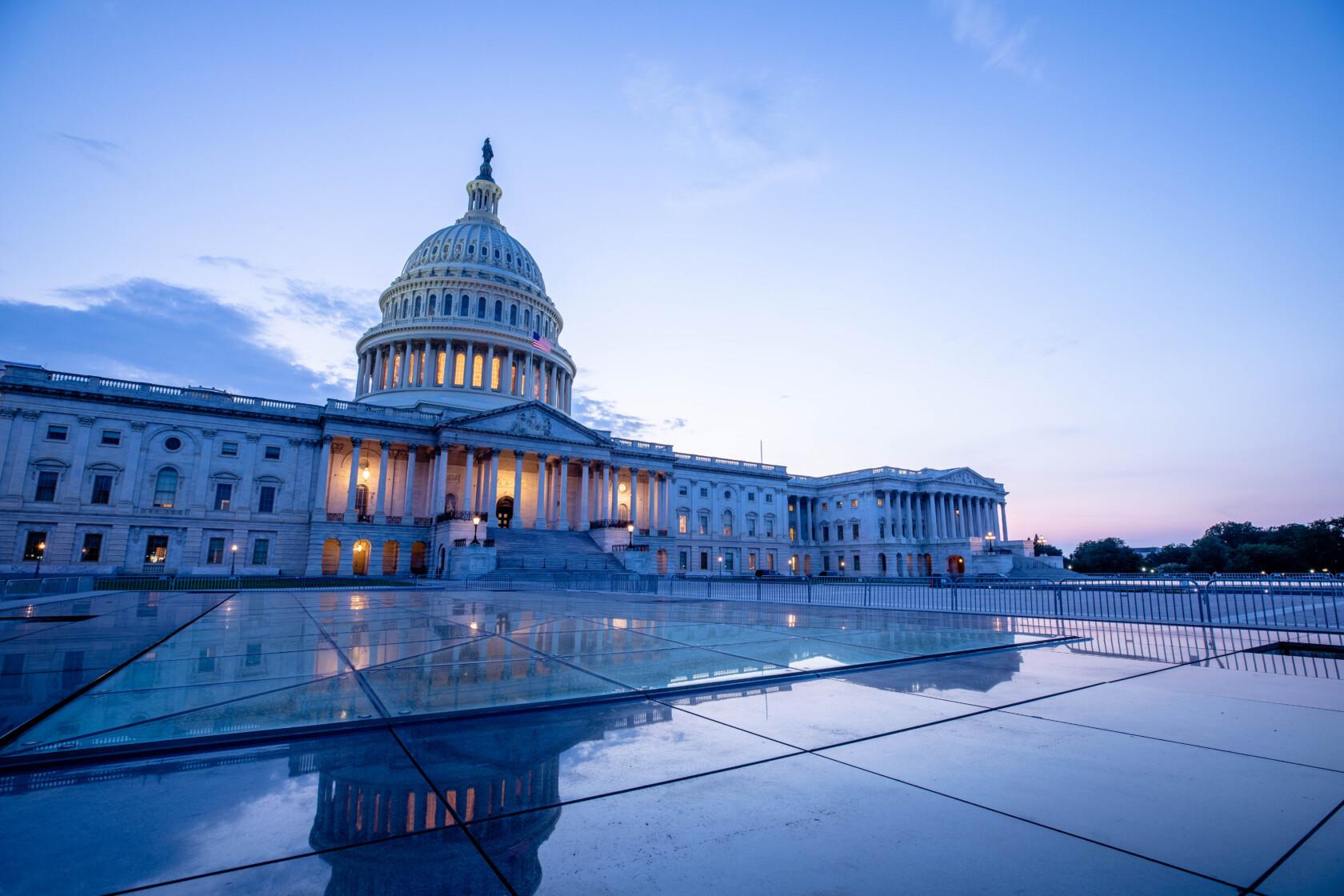 The United States Capitol building at dusk, illuminated with lights, stands under a cloudy sky with reflections on a wet surface in the foreground—an inspiring sight for those in corporate law offices nationwide.