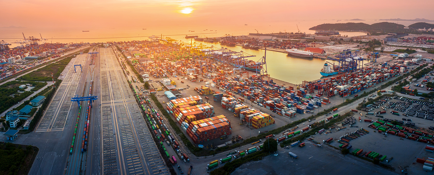 Aerial view of a busy shipping port at sunset, showing rows of colorful shipping containers, cranes, trains, and docked ships—much like the organized efficiency found in top law offices specializing in intellectual property law.