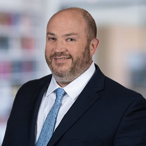 A man in a navy suit, white shirt, and light blue tie is smiling, posing in front of a blurred corporate law office, reflecting the professionalism of top lawyers in Chicago.