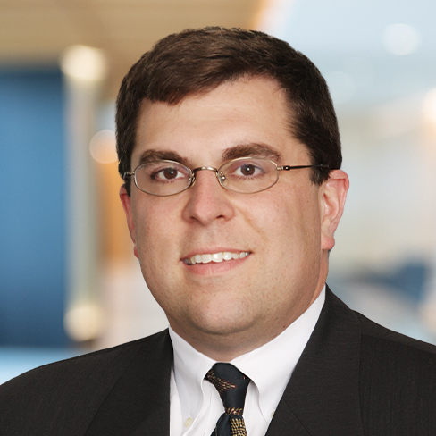A man wearing glasses, a dark suit, a white shirt, and a patterned tie stands posed in front of a blurred corporate law office, reflecting the professional environment of top lawyers in Chicago.