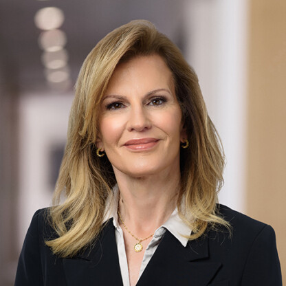 A woman with shoulder-length blonde hair, wearing a black blazer, white blouse, and gold jewelry, stands in a blurred indoor setting often seen in law offices specializing in litigation support.