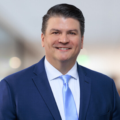 A man in a blue suit and light blue tie smiles at the camera, standing in a blurred indoor corporate law office setting.