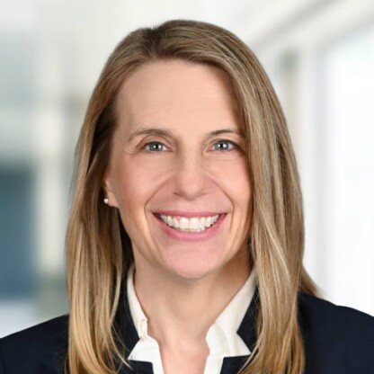 A woman with straight, shoulder-length blonde hair, wearing a dark blazer over a white collared shirt, smiles at the camera in a bright, blurred corporate law office—perfectly capturing the professional spirit of lawyers in Chicago.