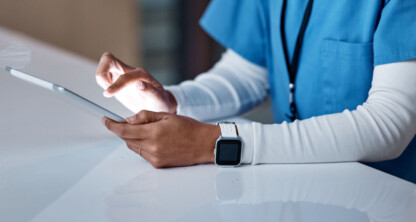A person in medical scrubs uses a tablet at a white desk, wearing a smartwatch and an ID badge. Their face is not visible, suggesting routine tech use in busy law offices or even a corporate law office environment.