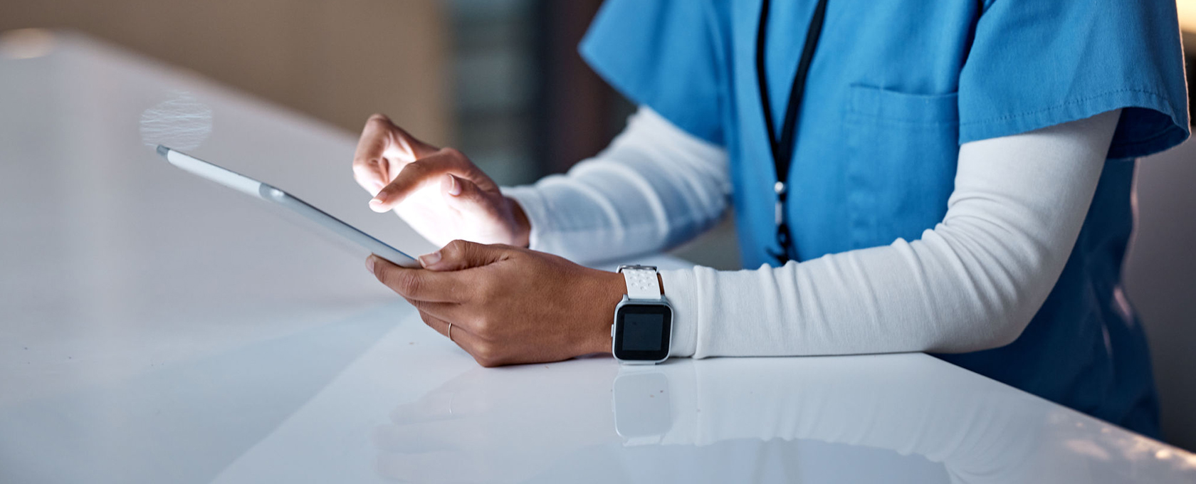 A person in medical scrubs uses a tablet at a white desk, wearing a smartwatch and an ID badge. Their face is not visible, suggesting routine tech use in busy law offices or even a corporate law office environment.