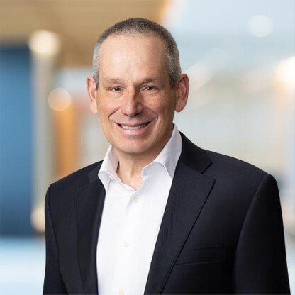 A middle-aged man in a suit and white shirt smiles at the camera, standing in a modern, blurred indoor setting typical of corporate law offices.