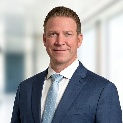 A man in a blue suit and light blue tie stands in front of a blurred corporate law office background, facing the camera and smiling slightly.