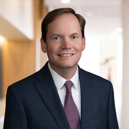 A man in a dark suit and red patterned tie stands indoors, smiling at the camera with a blurred corporate law office background—an image often seen among Chicago lawyers or those specializing in intellectual property law.