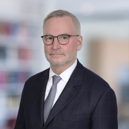 A man with short gray hair, glasses, and a beard, wearing a dark suit, white shirt, and tie, stands in front of a blurred office background, representing litigation support for lawyers in Chicago.