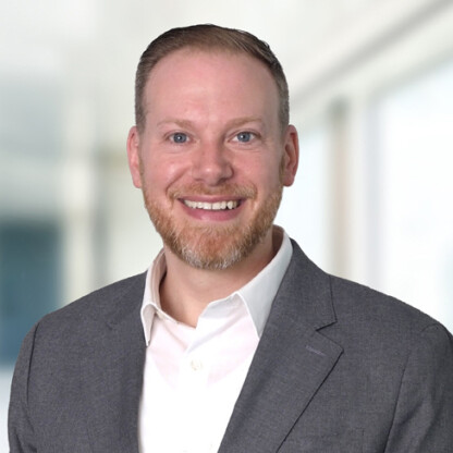 A man with short light hair, a beard, and light skin is wearing a gray suit jacket and white shirt, smiling at the camera against a blurred indoor background at one of Chicago's top corporate law offices.
